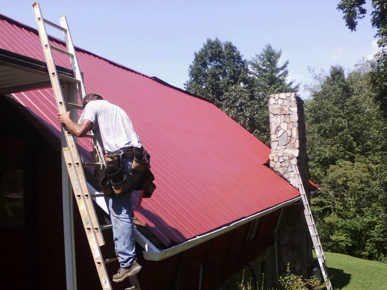 Roofer on a ladder beside a red metal roof and stone chimney — Soffit & Fascia Repair by Roofing Brothers in Ogdensburg